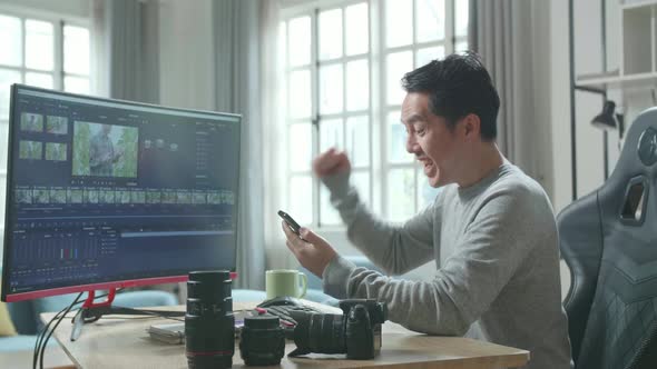 ໊Happy Asian Video Editor Man Looking At Mobile Phone While Using Computer Editing Video At Home
