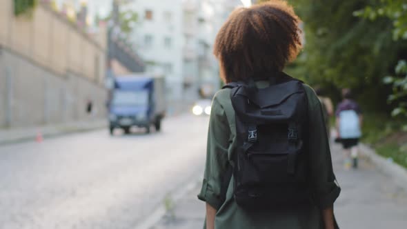 Back View Young African American Tourist with Afro Hair and Backpack Walking Alone Along Road