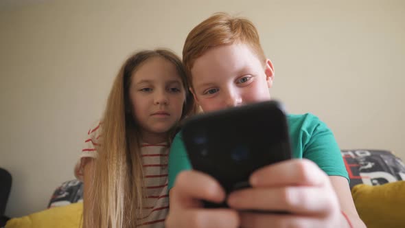 Close Up of Little Smiling Brother and Sister Playing the Games on Mobile Phone at Home. Happy Small