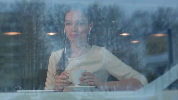 Smiling Young Woman at the Cafe with Headphones Listening To Music From Tablet