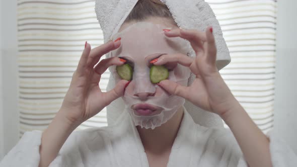 Attractive Young Woman Eating Cucumber with the Sheet Mask on Her Face in the Bathroom