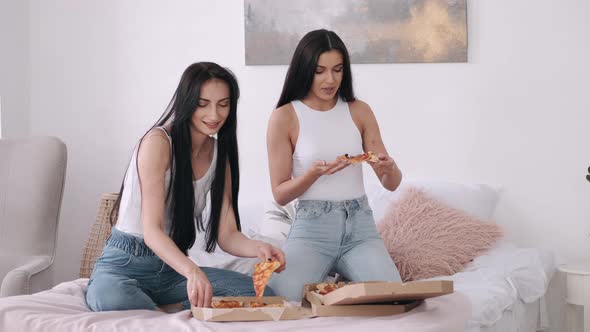 Two Dark Hair Pretty Ladies Are Sharing a Takeaway Pizza on the Bed