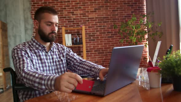 Disabled Hipster Man is Sitting in Wheelchair and Holding Credit Card and Using Laptop Side View