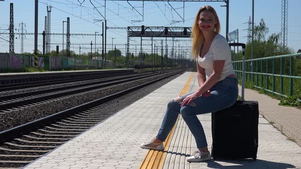 A Young Beautiful Woman Sits on a Suitcase on a Train Station Platform and Smiles at the Camera