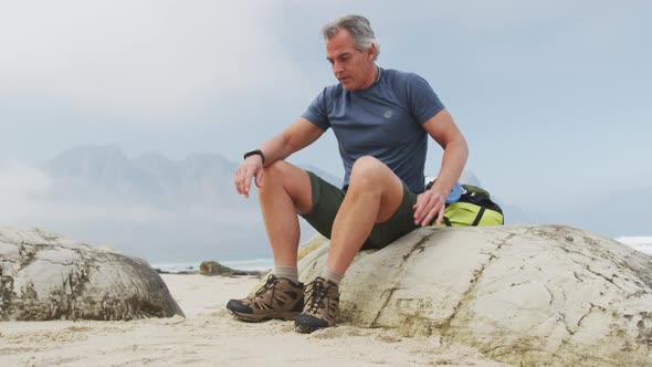 Tired senior hiker man with backpack using smartwatch while sitting on rock hiking on the beach.