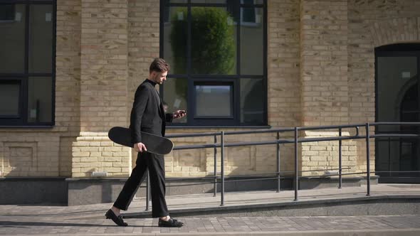 Side View Serious Successful Young Businessman Walking in Sunlight on City Street with Skateboard