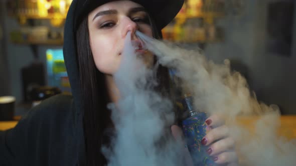 Young Pretty Woman in Cap Smoke an Electronic Cigarette at the Vape Shop. Hip-hop Style. Closeup.