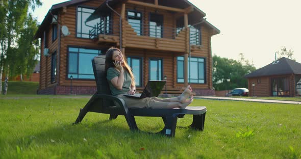 Young Happy Girl Sitting with Laptop in Lawn Chair at Backyard of Country House and Talking on