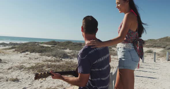 Happy caucasian couple sitting on the beach by the sea playin gguitar