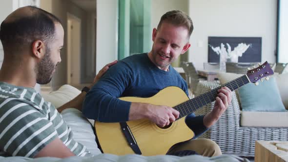 Multi ethnic gay male couple sitting on couch one playing guitar