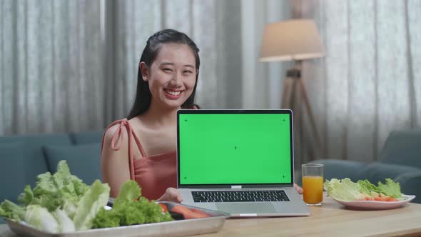 Smiling Asian Woman Showing Green Screen Laptop To Camera While Preparing Healthy Food At Home