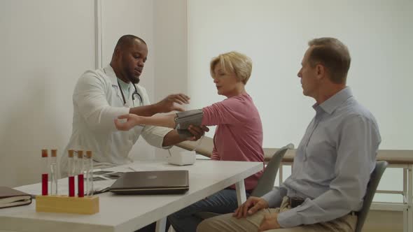 Black Doctor Checking Blood Pressure and Heartbeat of Middle Aged Woman Using Electronic Device