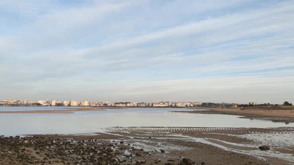 Shallow Riverside With Calm Water And Cityscape In Background Under Cloudy Sky. - Timelapse