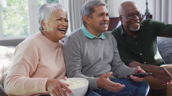 Happy biracial senior couple and african american friend watching tv with popcorn