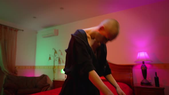 Side View of Emotional Male Queer Putting on Black Dress Standing in Bedroom with Red Lights