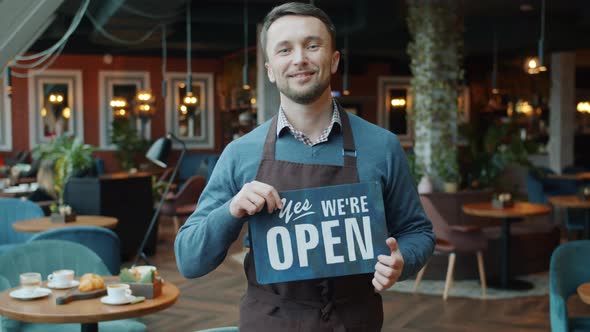 Slow Motion Portrait of Confident Young Man in Apron Holding Open Sign in Cafe