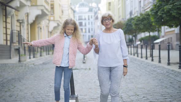 Smiling Beautiful Senior Woman Holding Hand of Cheerful Little Girl Walking Along Curb. Portrait of