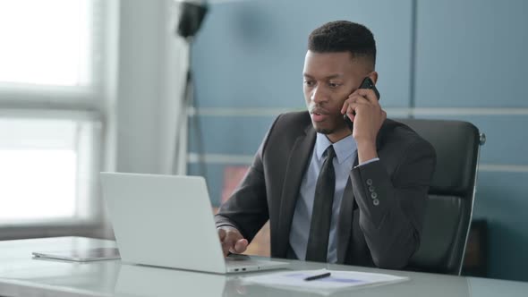 African Businessman Talking on Smartphone while using Laptop in Office