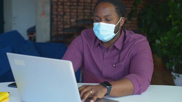 Concentrated AfricanAmerican Male Employee Wearing Medical Mask Using Laptop