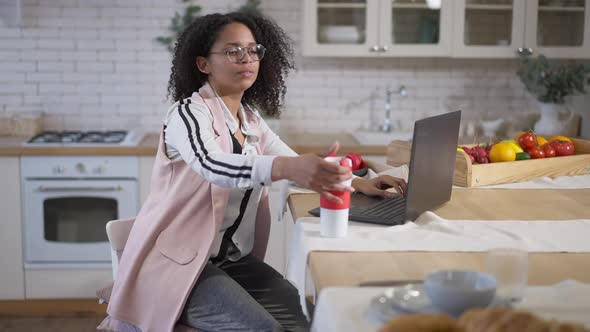 Concentrated Serious African American Woman Typing on Laptop Keyboard and Drinking Coffee From
