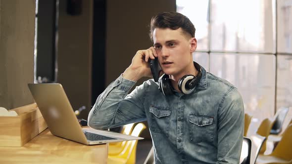 Attractive Male in His 20's in Denim Shirt with Wireless Headphones Around His Neck and Laptop