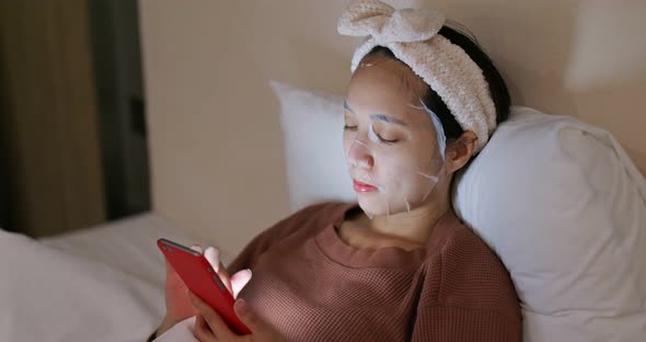 Woman use of mobile phone and apply the face mask on bed at night