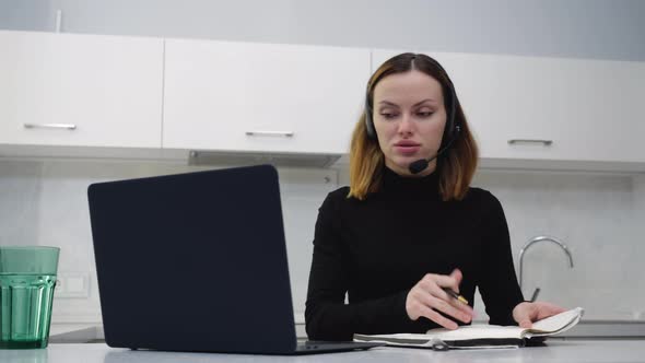 A Young Smart Girl Talking on the Headset Making Notes in a Notebook