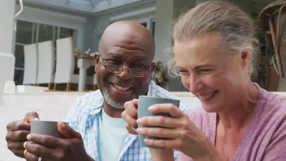 Happy senior diverse couple wearing shirts and drinking coffee in garden