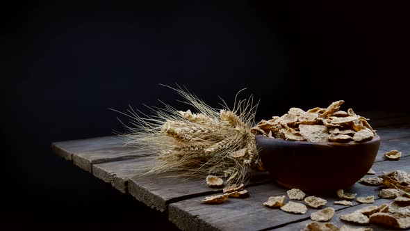 wheat flakes fall into a clay plate on a wooden table. Bowl of dry wheat flakes with ears.