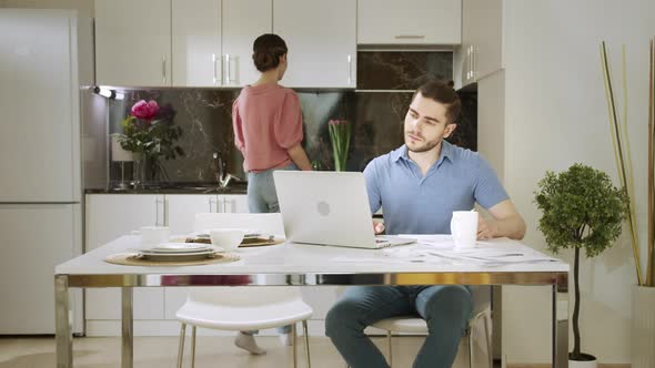 An Adult Man is Sitting and Working at Home While His Wife is Doing Household Chores