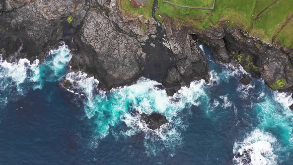 Aerial View of Waves Break on Rocks of Faroe Islands Cliffs in a Blue Ocean