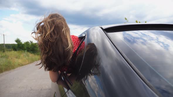 Brunette Girl Leaning Out of Car Window and Enjoying Trip While Riding Through Country Road. Young