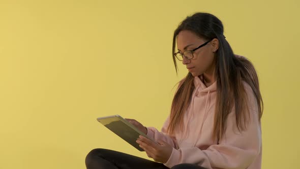 Black Woman in Eyeglasses Working on Tablet on Yellow Background.