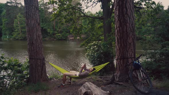Man Traveling on Bicycle Resting in Green Hammock Surfing Internet on Smartphone in Forest Near Lake