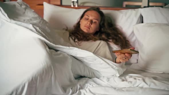 A Young Woman Lies Down in Bed with a Phone in Her Hands