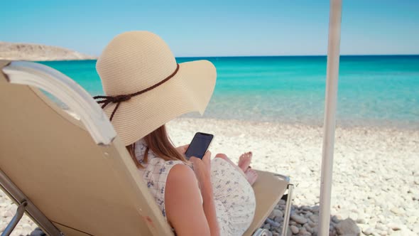 Lady in Hat Lying Uses Smartphone on Beach By Sea in Crete Grece