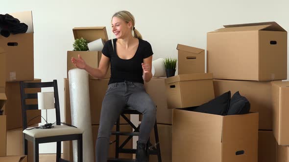 A Happy Moving Woman Sits on a Chair in an Empty Apartment and Celebrates, Surrounded By Boxes