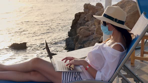 Young Woman in Protective Mask, Sun Hat and Summer Clothes, Working on Laptop, Sitting on Sun