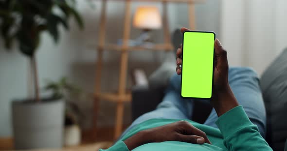 Young Man Lying on Couch with Modern Green Screen Smartphone in His Hand. Guy Watching Video While
