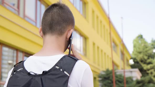 A Caucasian Teenage Boy Talks on a Smartphone  Rear Closeup From Below  a School in the Background