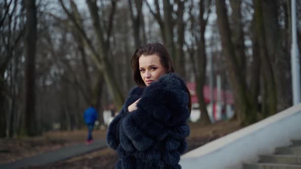 Young beautiful fashionable lady wearing stylish dark fur coat posing in park. Model looking aside. 