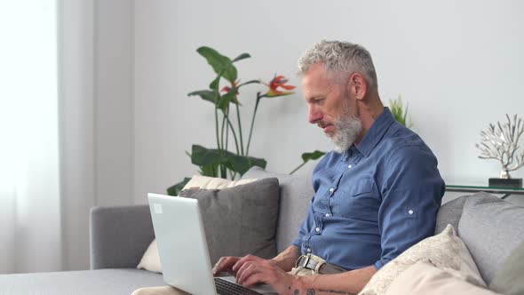 Middleaged Hipster Man in Smart Casual Shirt Using Laptop Sitting on the Couch at Home