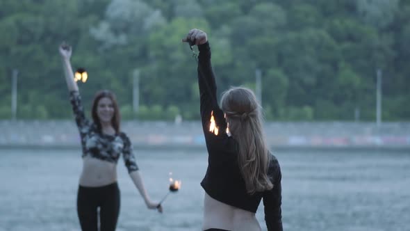 Two Young Women Performing a Show with Flame Balls Standing on the Riverbank