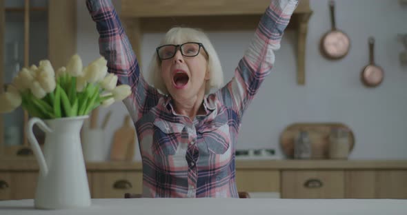 Energetic Elderly 70s Woman in Glasses Rising Hands Up Successfully, Sitting at Dining Table with