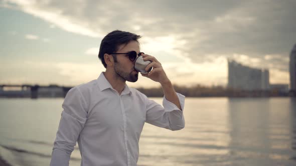 Businessman Drinking Tea Or Coffee From Steel Cup And Enjoys View. Man Relaxing On City Beach .