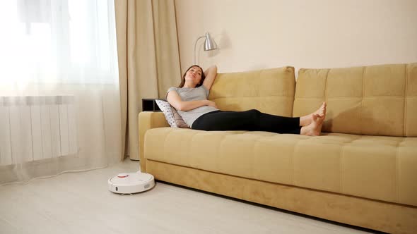 Happy Woman Lies on Sofa While Robot Cleaner Hoovers Floor