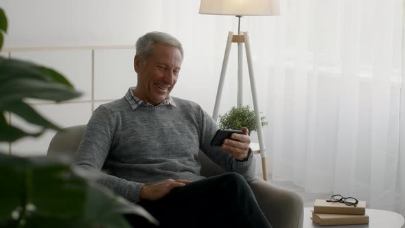 Smiling Senior Man Relaxing at Home with Smartphone Watching Funny Videos Online