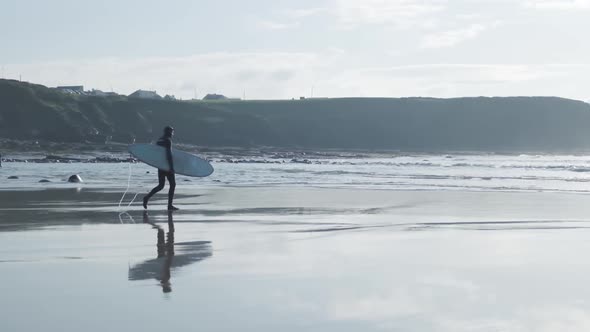 Medium wide shot of a surfer in a wet suit carrying his surfboard under his arm on a wet beach in Ir