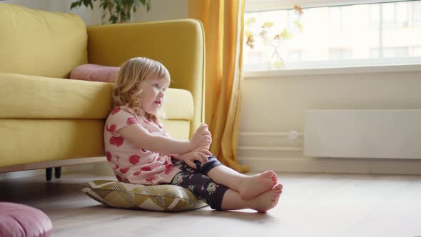 Little Cheerful Girl Child in Pink Dress Sits on Floor in Living Room Next to Yellow Sofa Eats