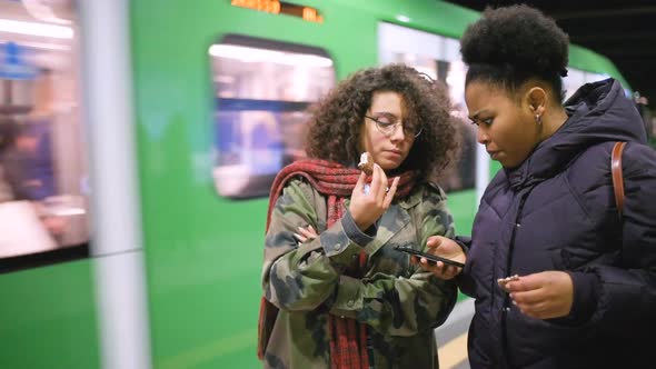 Two young multiethnic women in the underground eating snack using smart phone